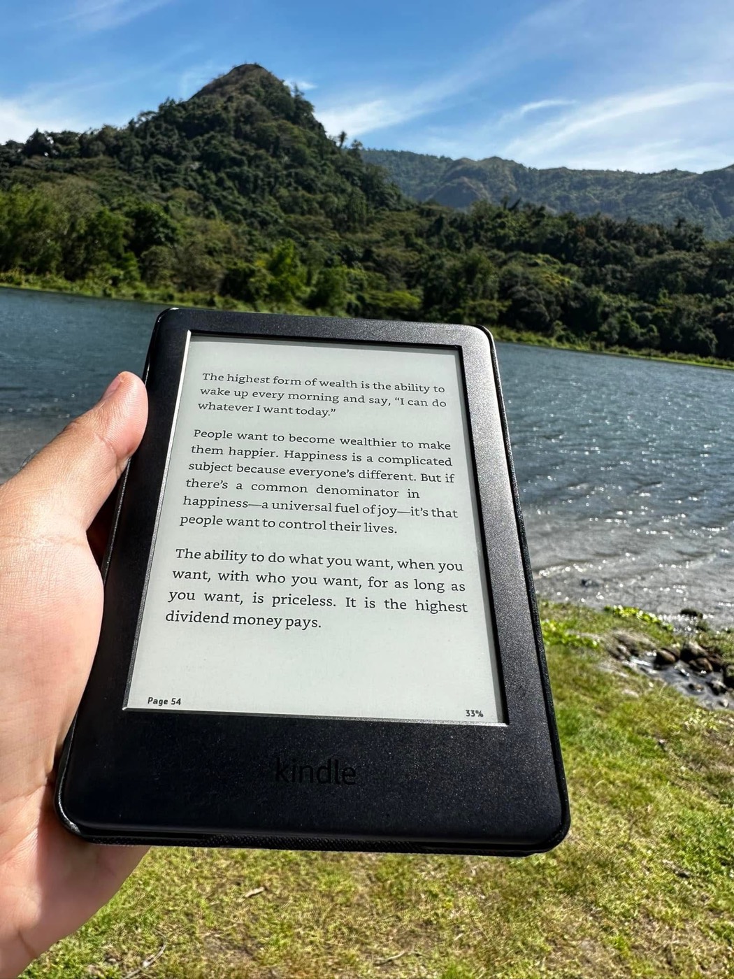 Kindle showing a quote about wealth, held up against a lake and mountain